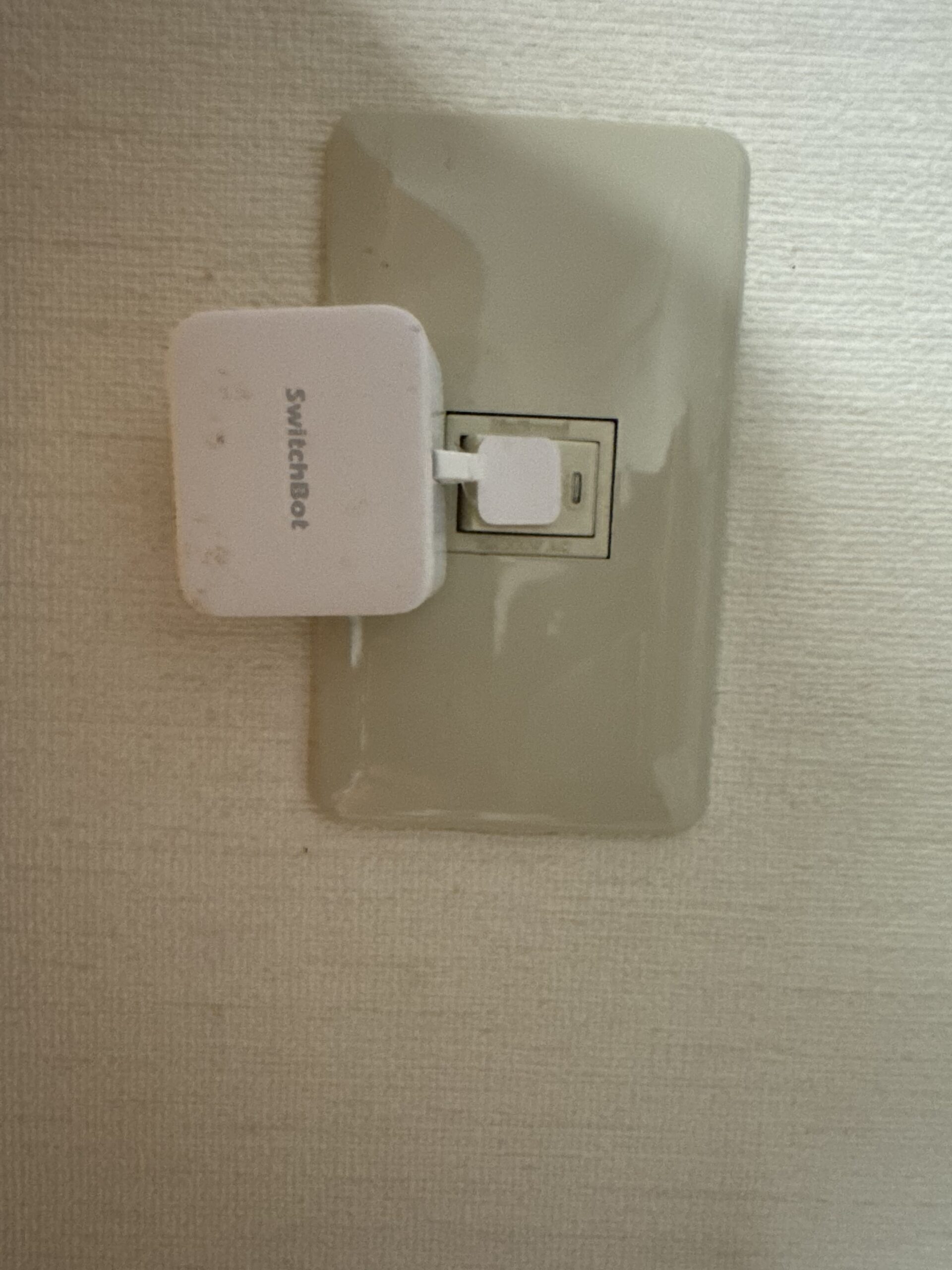 SwitchBot ボットを壁の照明スイッチに取り付けた写真。白いコンパクトな本体がスイッチを自動で押す仕組み